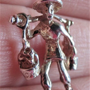 May include: Silver charm of a person carrying a basket on their shoulder. The person is wearing a wide-brimmed hat and traditional clothing.