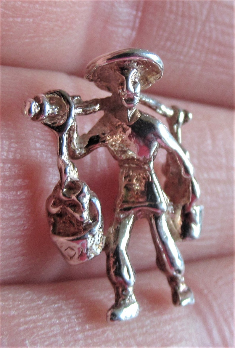 May include: Silver charm of a person carrying a basket on their shoulder. The person is wearing a wide-brimmed hat and traditional clothing.