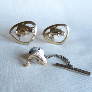 May include: A set of gold-toned fish-themed accessories, including a pair of cufflinks and a tie tack. The cufflinks feature a fish within a heart-shaped frame. The tie tack has a fish design with a chain and bar.