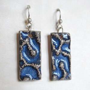 Puede incluir: Un par de pendientes colgantes rectangulares. Cada pendiente presenta un patrón floral azul y plateado sobre una baldosa de cerámica rectangular. Los pendientes cuelgan de alambres plateados.