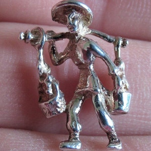 May include: Silver charm of a person carrying two buckets on a stick, wearing a hat and traditional clothing.