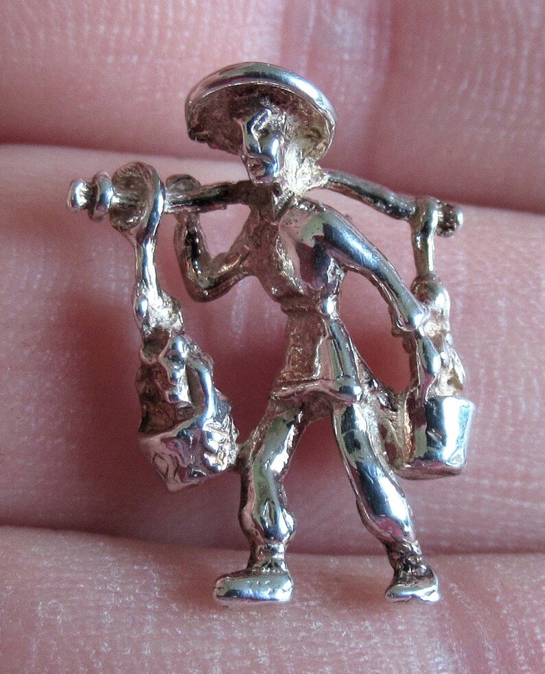 May include: Silver charm of a person carrying two buckets on a stick, wearing a hat and traditional clothing.