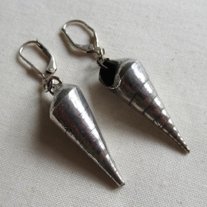 Peut inclure: Une paire de boucles d'oreilles pendantes argentées en forme de cône rappelant un coquillage, avec des stries horizontales et une finition métallique lisse. Chaque boucle est fixée à une attache dormeuse argentée.