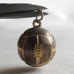 May include: A small, round, metallic charm shaped like a football, featuring a textured, dark-toned surface with raised gold-coloured lines and laces. The charm includes a small circular jump ring at the top for attachment to a chain or bracelet.