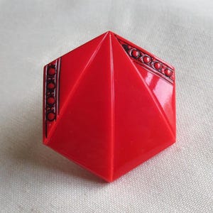 May include: A red geometric brooch with a faceted design. The brooch has a hexagonal shape with a pyramid-like structure. Darker red decorative accents are on the edges, adding contrast to the bright red colour. The brooch is set against a white background.