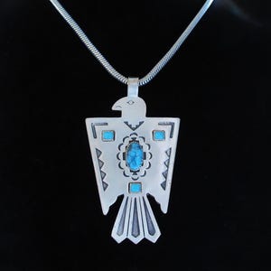 Puede incluir: Collar con colgante plateado con diseño de ave estilizada. El colgante tiene una piedra central ovalada de turquesa y pequeños detalles turquesas. El collar tiene una cadena plateada, sobre fondo negro.