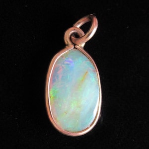 May include: An oval-shaped opal pendant set in a smooth, polished metal bezel. The gemstone displays a milky white base color with iridescent flashes of green, blue, and pink light. A small, circular metal jump ring is attached to the top.
