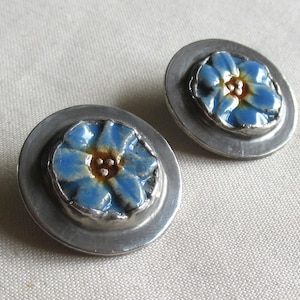 Könnte beinhalten: Ein Paar silberfarbene Ohrstecker. Jeder Ohrring hat ein blaues Blumenmuster mit gelbem Zentrum, das in einem kreisförmigen Rahmen eingefasst ist. Die Ohrringe werden auf einem neutralfarbenen Stoff präsentiert.