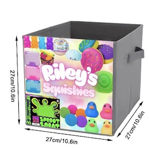 Puede incluir: Un contenedor de almacenamiento de tela gris con un gráfico colorido de juguetes blandos, que incluyen donas, patos y ositos de goma. El contenedor mide 27 cm cuadrados y presenta el texto "Riley's Squishies". Tiene dos asas grises.
