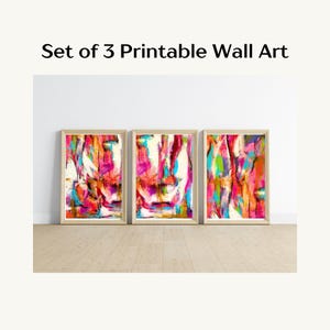 May include: Set of 3 framed abstract art prints. Each print features a vibrant mix of colors including pink, orange, blue, and white. The artwork is displayed in a light-colored wooden frame. The text "Set of 3 Printable Wall Art" is at the top.