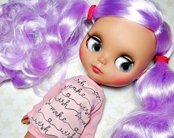 May include: A doll with a pale complexion, large eyes, and a pouty expression. The doll has long, wavy, lavender hair styled in pigtails secured with red bands. It wears a pink sweater with the words "make a wish" and star graphics.