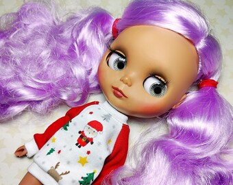 May include: A doll with large eyes and long, lavender hair styled in pigtails. The doll wears a red and white Christmas-themed sweater with a Santa Claus graphic, a reindeer, and Christmas trees.