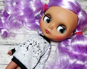 May include: A doll with a fair complexion, large gray eyes, and lavender pigtails. The doll is wearing a white sweater with black and blue paint splatters and black trim. The doll's hair is styled in two high pigtails, secured with red bands.