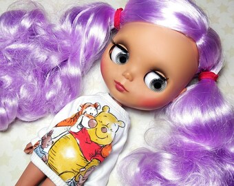 May include: A doll with a light skin tone, large gray eyes, and purple hair styled in pigtails. The doll wears a white long-sleeved shirt featuring a graphic of Winnie the Pooh and Tigger. The doll's hair is secured with red hair ties.