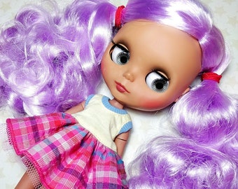 May include: A doll with long, curly, lavender hair wearing a pink and white plaid dress with a white and blue striped shirt underneath. The doll has brown eyes and a light brown complexion.