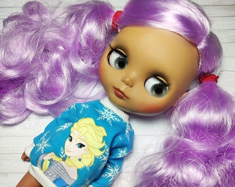 May include: A doll with long, purple hair wearing a blue sweater with a snowflake pattern and a picture of Elsa from Frozen. The doll has brown skin and blue eyes.