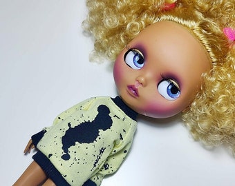 May include: A Blythe doll with large blue eyes and blonde curly hair. The doll wears a light yellow sweater with a black splatter design and a black collar and cuffs. The doll's face is painted with pink blush and purple eyeshadow.