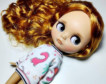 May include: A Blythe doll with large blue eyes and auburn hair. The doll wears a white sweatshirt with a pink dinosaur and heart pattern. The sweatshirt has a pink, white, and navy blue striped trim.