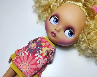 May include: A doll with a fair complexion, blue eyes, and blonde curly hair. The doll wears a pink floral print sweater with a yellow collar and cuffs. The doll's face has pink blush and detailed eye makeup.