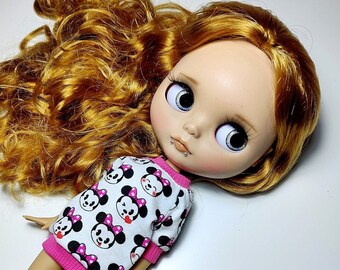 May include: A doll with large blue eyes, light skin, and auburn hair. The doll wears a white and pink long-sleeved shirt with a pattern of Minnie Mouse faces. The doll has a lip piercing.