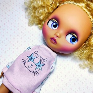 May include: A doll head with large blue eyes and blonde curly hair. The doll is wearing a pink sweatshirt with a cat face design and star-shaped sunglasses. The doll's face has pink blush and purple eyeshadow.