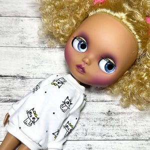 May include: A doll with a painted face, blue eyes, and blonde curly hair. The doll is wearing a white sweatshirt with black cat illustrations and black dots. The doll's face has pink blush and purple eyeshadow.