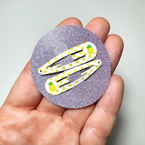 Puede incluir: Dos pinzas para el pelo blancas con un patrón de piña amarilla sobre un fondo de purpurina morado.