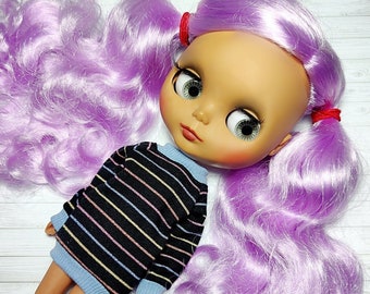May include: A doll with a large head, big eyes, and long, wavy lavender hair styled in pigtails with red ribbons. The doll wears a black sweater with horizontal stripes in various colors and a light blue collar and cuffs. The doll's skin is pale with rosy cheeks.