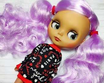 May include: A doll with a light skin tone, green eyes, and long, wavy lavender hair styled in pigtails. The doll is wearing a black and red sweater with the words "Paris" and "New York" printed on it. The doll's legs are visible.