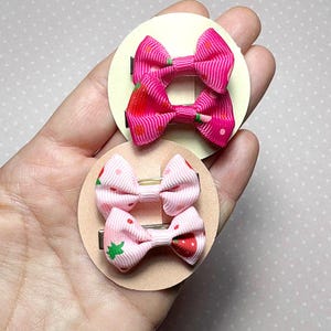 Op de afbeelding: Twee sets roze haarstrikken met een aardbeienthema. Elke strik is bevestigd aan een metalen clip en gemonteerd op een ronde, lichtbruine achtergrond. De strikken hebben een geribbelde textuur en aardbeienontwerpen, met rode en groene accenten.