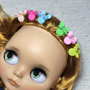 Può includere: Una bambola con i capelli biondi che indossa una varietà di fermagli per capelli in plastica colorati a forma di orecchie di Topolino. La bambola ha gli occhi azzurri e una carnagione chiara.