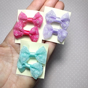 Puede incluir: Colección de pequeños lazos para el cabello de tela en tonos rosa, morado y turquesa. Cada lazo está adornado con un delicado estampado floral blanco. Los lazos están sujetos a pequeñas tarjetas rectangulares.