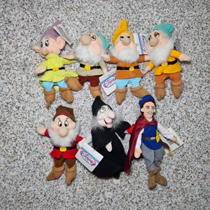 May include: A collection of seven plush dolls featuring characters from Snow White, including six dwarfs with white beards, colourful tunics, and pointed hats, one witch in a black robe, and one prince in a blue tunic and red cape, all with Disney tags.