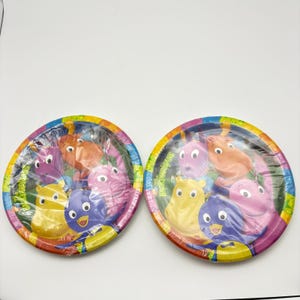 May include: Two round paper plates featuring The Backyardigans characters including Pablo, Tyrone, Uniqua, Tasha, and Austin. The plates have a colorful border and are wrapped in clear plastic. The text The Backyardigans is printed on the side.