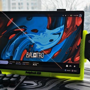 Op de afbeelding: Een tablet met het spel "BALATRO" in een neon groene standaard met aangesloten gamecontrollers. Het scherm toont een blauw en rood abstract ontwerp. Een toetsenbord is zichtbaar op de voorgrond.