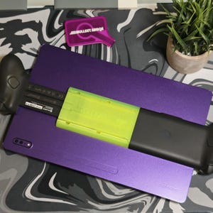 May include: A purple Samsung phone case with a black controller and a neon green accessory. The phone case is rectangular and has the Samsung logo at the bottom. A small, pink, rectangular object with text is also visible.
