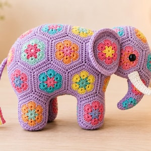 Puede incluir: Una figura de elefante de ganchillo construida con motivos hexagonales de color lavanda con centros florales coloridos en tonos amarillo, rosa y turquesa. El elefante tiene colmillos blancos, un ojo negro y una borla multicolor en la cola.