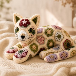 May include: A crocheted cat figurine constructed from numerous small, multi-colored floral hexagon motifs. The cat features large dark green eyes, a black embroidered nose and whiskers, and a cream-colored base yarn with accents of burgundy, olive green, and soft lavender.