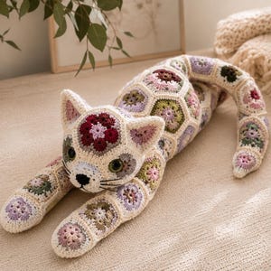 Könnte beinhalten: Ein Stoffkatzenspielzeug, das aus zahlreichen gehäkelten sechseckigen Motiven in den Farben Creme, Altrosa, Salbeigrün, Lavendel und Dunkelburgund besteht. Die Katzenfigur hat dunkle Kunststoffaugen, eine gestickte schwarze Nase und Schnurrhaare.