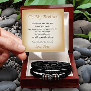 Puede incluir: Una pulsera de cuero trenzado negro con detalles metálicos y el texto "love you forever" dentro de una caja de regalo de madera oscura. Se incluye una tarjeta con el texto: "To My Brother, Thank you for being there when I need you most, I am blessed to have you appear in my life. Our paths may change, but the bond between us will always be strong. Love, Sister."