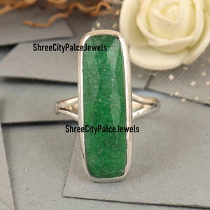 May include: A silver ring featuring a long, rectangular, faceted green gemstone set in a smooth metal bezel. The text ShreeCityPalceJewels is visible on the surface of the stone. The band has a split design that connects to the central setting.