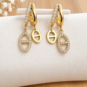 May include: A pair of gold-toned hoop earrings featuring a row of clear crystals along the front edge. Each earring suspends two oval-shaped charms, one encrusted with clear crystals and the other a plain polished gold-toned oval with a horizontal bar.