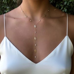 Peut inclure: Collier doré avec un design en forme de Y. Le collier présente de petits pendentifs ovales et de minuscules accents étincelants. Le collier est porté sur un haut blanc, créant un contraste qui met en valeur le bijou.