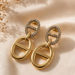 May include: A pair of gold-toned drop earrings featuring a two-part design. The top section is an oval link encrusted with small clear crystals, while the bottom section is a smooth, polished gold-toned oval link with a horizontal bar across the centre.