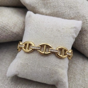 Op de afbeelding: Een goudkleurige armband met een schakelontwerp, bezet met heldere kristallen. De armband wordt gepresenteerd op een klein, vierkant, wit kussen, tegen een neutrale achtergrond.