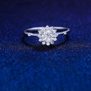 Peut inclure: Une bague en argent avec une pierre centrale ronde et transparente entourée de petites pierres en forme de fleur. La bague a de petites pierres de chaque côté de la bande, sur fond bleu foncé.