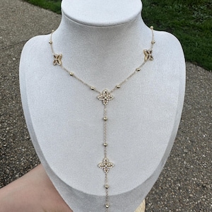 Op de afbeelding: Een goudkleurige ketting met een Y-vormige hanger. De ketting heeft vierbladige bloemvormige hangers en kleine ronde kralen. De ketting wordt getoond op een witte mannequin tegen een vage buitenachtergrond.