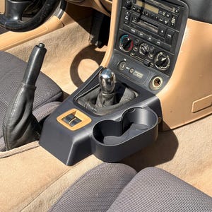 May include: A black plastic centre console cup holder insert installed in a vehicle interior with tan carpeting and upholstery. The unit features two circular cup slots, a gear shift lever with a metallic knob, and a black leather parking brake handle.
