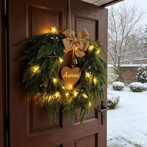Op de afbeelding: Een ronde kerstkrans van donkergroene dennentakken, versierd met een grote strik van jute en kleine metalen belletjes. In het midden hangt een houten hartvormig ornament met de naam Aurora in goud gegraveerd, verlicht door warme sterlampjes.