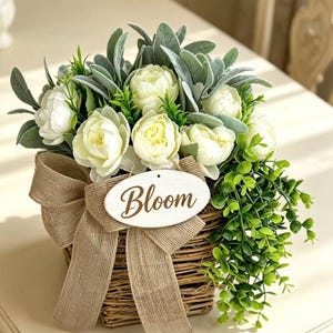 Può includere: Un cesto in vimini intrecciato riempito con fiori di peonia bianchi, fogliame verde e foglie color salvia. Un nastro di iuta color marrone chiaro è legato sul davanti, con un cartello ovale in legno bianco con la parola Bloom in corsivo.
